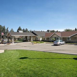 A single-story building with a driveway and a parked car, surrounded by trees and a well-maintained lawn under a clear blue sky.