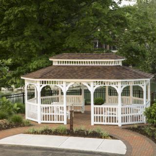 A charming white wooden gazebo in a garden setting, surrounded by trees and landscaping, with a paved walkway leading up to it.
