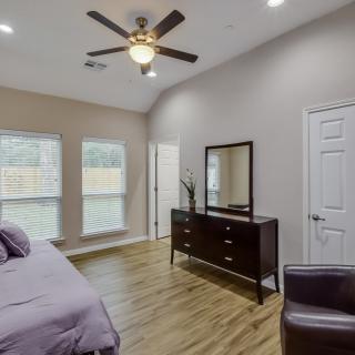 A modern bedroom with wooden flooring, featuring a bed, dresser with mirror, ceiling fan, and large windows.
