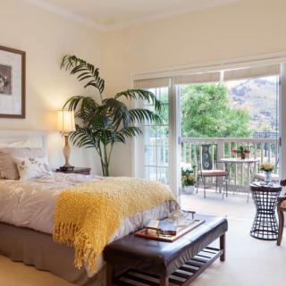 A cozy and elegantly decorated bedroom with a large window leading to a balcony, allowing natural light to fill the space.