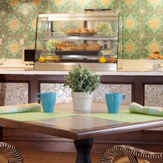 A cozy cafe interior with a wooden table set for two, featuring blue cups and a potted plant. The background showcases a display counter and elegant green patterned wallpaper.
