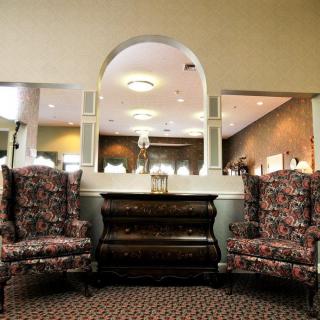 A cozy interior scene featuring a vintage piano flanked by two patterned armchairs under a decorative archway and mirror.