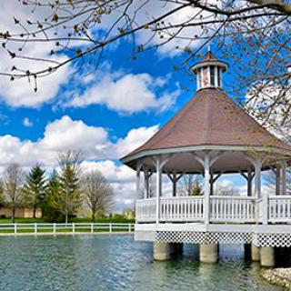 A scenic gazebo over a pond surrounded by trees and a white fence on a bright spring day.