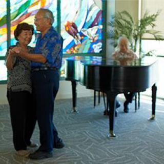 A couple dances joyfully near a pianist in front of colorful stained glass.
