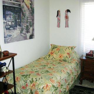 A small, cozy bedroom with a floral bedspread, wall art, and a nightstand.