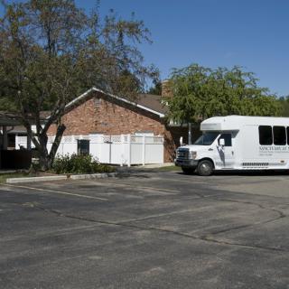A white bus parked in front of a suburban house with a parking lot and trees on a clear day.