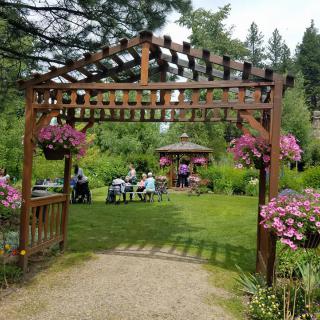 A picturesque garden setting with a wooden pergola, vibrant flowers, and a group of people gathered near a gazebo.