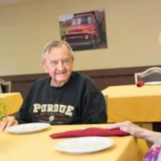 An elderly person wearing a Purdue sweatshirt sits at a dining table with others.