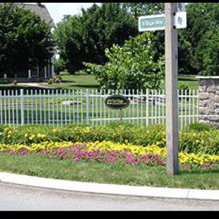A street corner featuring a white fence, vibrant flower beds, and lush greenery.
