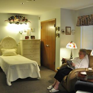 An elderly woman sitting and reading in a cozy bedroom with a lamp.