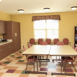 A small meeting room with tables and chairs, featuring a kitchenette and large window.