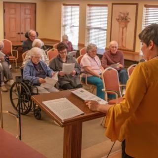 A woman presents to a group of elderly individuals gathered in a community room.