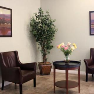 A cozy waiting room with leather chairs, a round table adorned with flowers, a potted plant in the corner, and framed paintings on the wall.