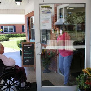 A senior person sitting in a wheelchair while another individual approaches the door of a care facility.