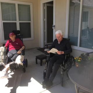 Two elderly individuals enjoying a sunny day on the patio, one reading a book.