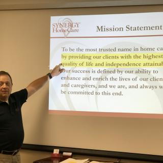 A man presents a mission statement on a projector screen in an office setting.
