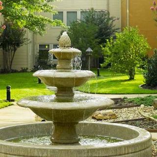 A multi-tiered fountain in a garden setting with lush greenery and a building in the background.