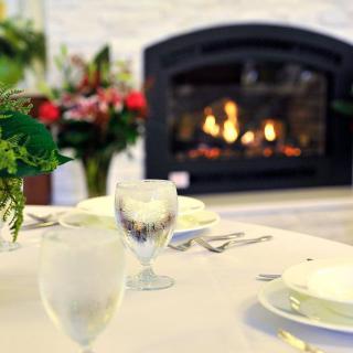 A cozy dining setup featuring a table elegantly set with glassware and surrounded by floral arrangements, with a warm fireplace glowing in the background.
