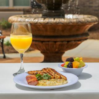A delightful breakfast featuring grilled chicken with herbs, a fresh fruit bowl, and a glass of orange juice served at an outdoor table with a fountain backdrop.