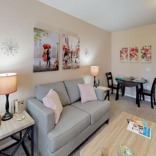 A cozy living room with modern decor featuring a gray sofa, artistic wall pictures, ambient lighting from lamps, and a dining area.