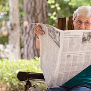 An elderly person sits on a bench outdoors, reading a newspaper.