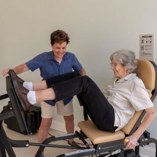 An elderly woman exercises on a leg press machine with the assistance of a trainer.