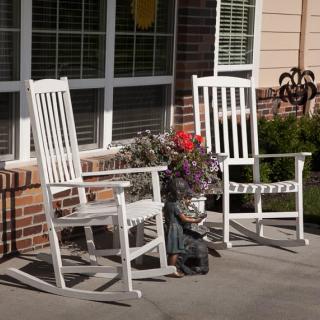 Two white rocking chairs on a sunny porch with flowers and plants.