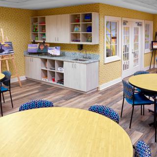 A bright art studio with round tables, chairs, and easels set against yellow walls and a hardwood floor.