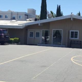 A small building with a parking lot and tall trees in the background on a sunny day.