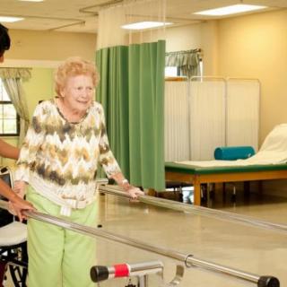 An elderly woman receives support during a physical therapy session in a healthcare facility.