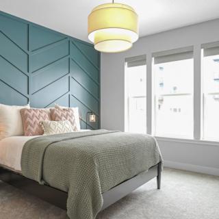 A modern bedroom with a blue accent wall, featuring a neatly made bed with decorative pillows and a green blanket. Ample natural light streams through large windows.