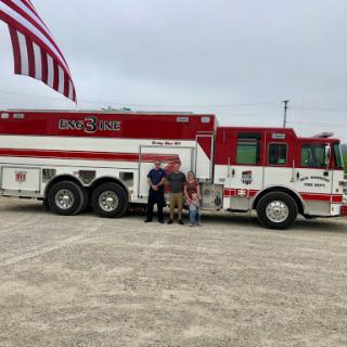 A group posing in front of a fire truck, with an American flag waving in the background.