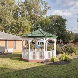 A quaint gazebo is surrounded by a well-maintained garden and lawn, with trees in the background on a sunny day.