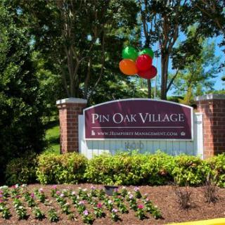 Entrance sign to Pin Oak Village with balloons and well-maintained landscaping.