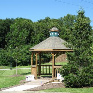 A wooden gazebo surrounded by greenery in a park setting with benches and a pathway.