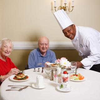 An elderly couple enjoying a meal served by a chef in a restaurant setting.