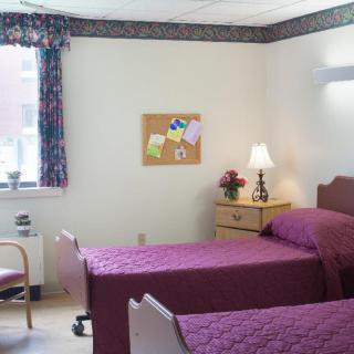 A hospital room with twin beds covered in purple bedspreads, featuring a nightstand with a lamp and a window with curtains.