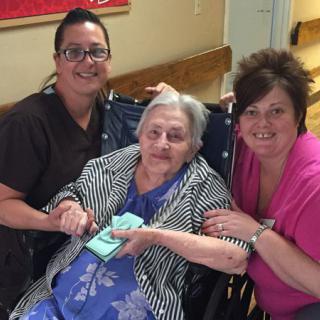 An elderly woman smiling while sitting in a wheelchair, accompanied by two caretakers indoors.