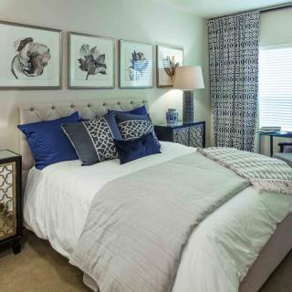 A modern bedroom with stylish decor, featuring a bed adorned with blue and patterned pillows, framed artwork on the wall, and elegant nightstands.