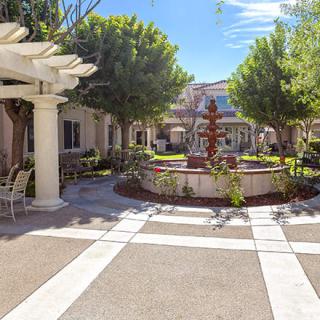 A sunny courtyard with a central fountain, surrounded by trees and outdoor seating under a pergola.