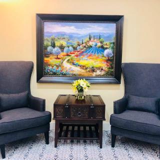A cozy living room setting with two chairs, an ornate table with flowers, and a colorful landscape painting.