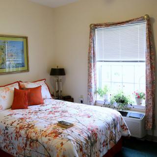 A cozy bedroom with floral bedding, decorated with plants on the windowsill and artwork on the wall.