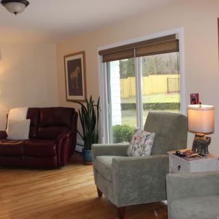 A cozy living room with armchairs and a recliner, featuring a lamp on a side table and a large window overlooking the yard.