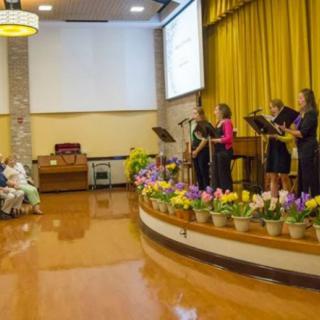 A choir performs on stage in a church, surrounded by colorful flowers, while an audience listens attentively.