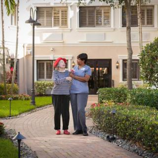 A caregiver assisting an elderly person on a walk in the garden of a healthcare facility.