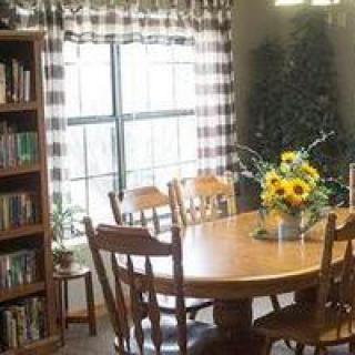 A cozy dining room with a wooden table, chairs, and a bookshelf by the window adorned with checkered curtains.