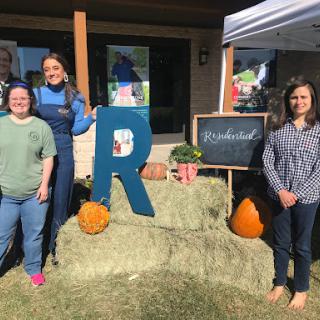 A cheerful outdoor gathering featuring people posing with colorful pumpkins and a large letter 'R' on a sunny day.