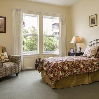 A cozy bedroom with a floral bedspread, armchair, and large windows.