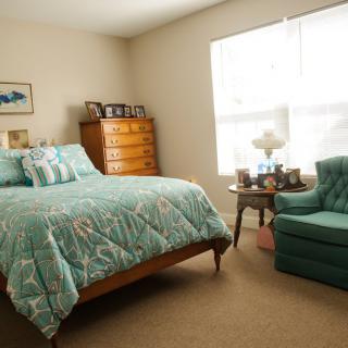 A cozy bedroom with teal bedding, an armchair, and a wooden dresser beside a large window.