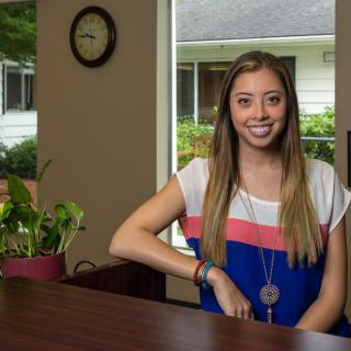 A woman smiling while sitting at a desk in an office with a plant and clock visible.
